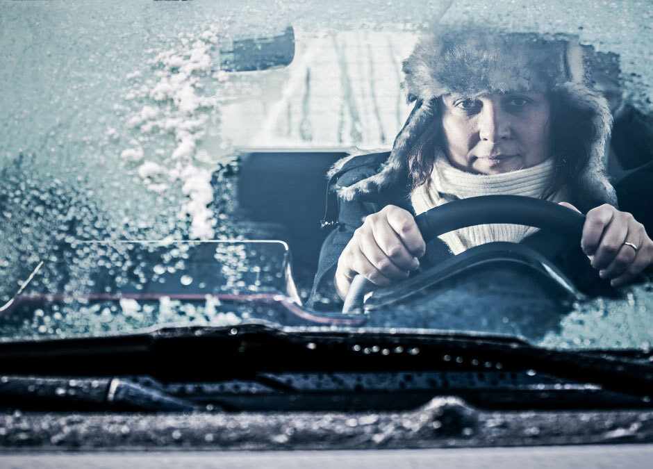 A bundled driver wearing a fur-lined hat and thick scarf grips the steering wheel from inside a vehicle, viewed through a heavily frosted and icy windshield
