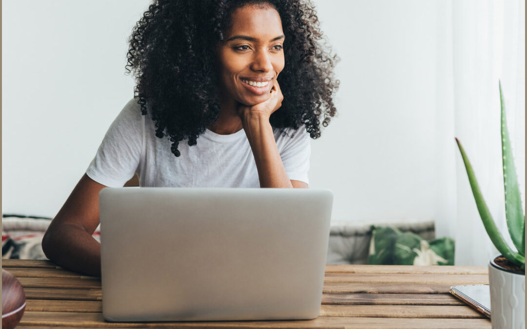 A smiling young woman with curly hair rests her chin on her hand while working on a laptop at a wooden desk, with an aloe vera plant nearby - www.koinsurance.com